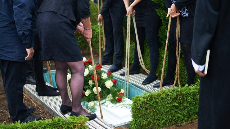 Teilnehmer einer Trauerfeier halten Seile, um Blumenschmuck auf einen Sarg in einer Grabstelle abzusenken.