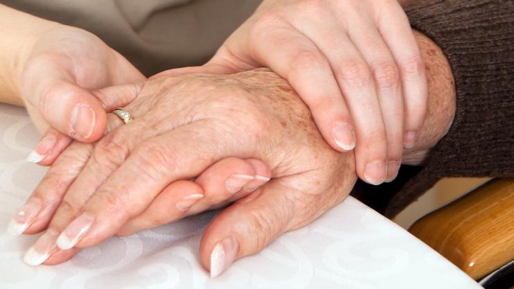 Zwei Hände, eine ältere und eine jüngere, liegen auf einem Tisch, wobei die jüngere Hand die ältere sanft umschließt.