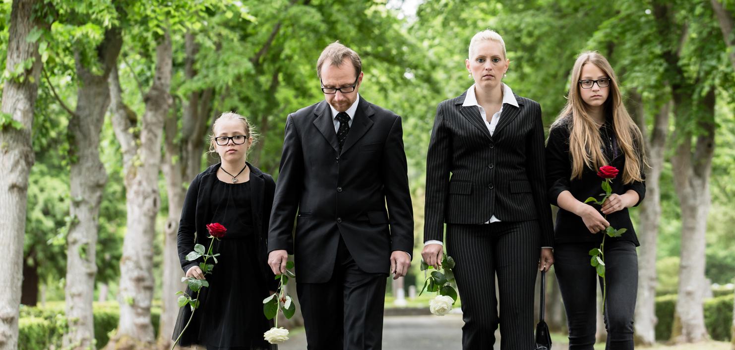 Vier Trauernde in Schwarz gehen auf einem gepflasterten Weg, halten Rosen in den Händen und sind von Bäumen umgeben.